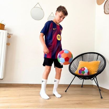 A young boy wearing a FC Barcelona soccer jersey, black shorts, and white socks stands in a room, holding a colorful House Ball next to a black chair with a yellow pillow.